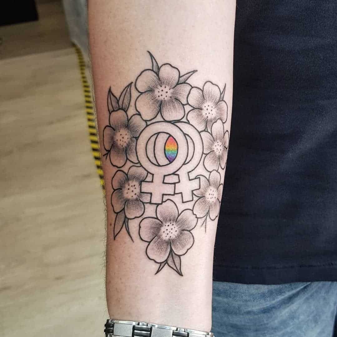 Tattoo op onderarm met 2 vrouwelijke tekens en een regenboog ertussen. Omringd door bloemen. Geplaatst bij Inksane tattoo en piercing.